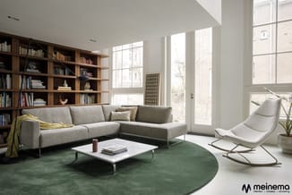 Moderne woonkamer met grijze hoekbank, witte schommelfauteuil, groen tapijt, witte coffeetable, houten boekenkasten en grote