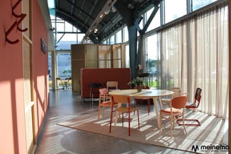 Moderne kantoorruimte met ronde houten tafel, oranje stoelen, rode loungeelementen, grote ramen met gordijnen en stalen const