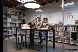 Twee personen bij zwarte werktafel in modern kantoor met witte kasten, grijze betonvloer en ronde plafondlamp.