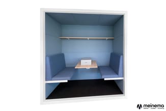 Blauwe belcel met zitbanken aan weerszijden, houten tafel met laptop, wandplank en zwarte mat op vloer.