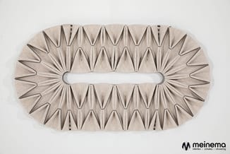 Beige akoestisch paneel met driehoekige vouwen en centrale opening, gezien van voren tegen witte wand.