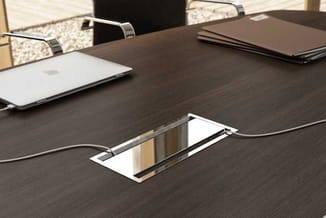 Donkere vergadertafel met ingebouwde witte kabelmanager, laptop en documenten, stoelen op achtergrond.