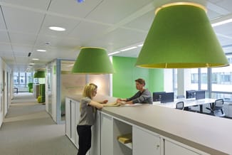 Moderne receptie met twee groene hanglampen met gele binnenrand, witte werkbank, twee vrouwen, kantoorruimte met groene wand