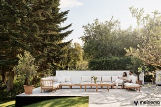 Houten loungeset met beige kussens op terras, omgeven door bomen en groen, twee personen zittend, meinema-logo rechtsonder.