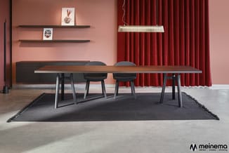Lange vergadertafel met houten blad en zwarte metalen poten, vier grijze stoelen, zwart tapijt, roze muren, rode gordijnen, w