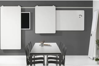 Vergaderruimte met grijze wanden, witte whiteboarden, zwarte stoelen rond tafel, flipchart en kantoormaterialen.