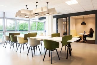 Modern kantoor met witte vergadertafel, groene en gele stoelen met zwart metalen frame, houten akoestische hanglampjes, raamp