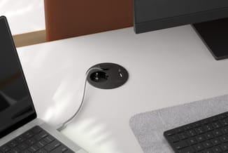 Zwart-witte desk power unit met stopcontact en USB-poort, geïntegreerd in wit bureau tussen toetsenborden en monitoren.