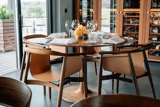 Ronde houten vergadertafel met 4 leren stoelen in moderne eetkamer met grote ramen, glazen wijnglaten en bloemencenterstuk.