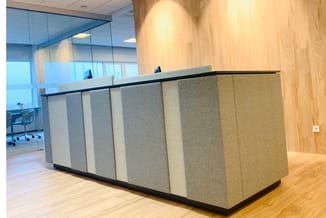 Receptiebalie met grijze en beige stoffen panelen, zwart werkblad en glazen wand in moderne kantoorruimte.
