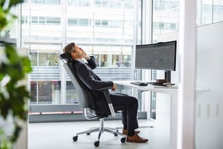 Zakenman in donker pak zit ontspannen in zwarte bureaustoel aan witte bureau met monitor, uitzicht op kantoorgebouwen.