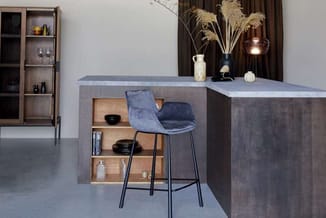 Moderne keukeneiland met betonnen afwerking, houten open kasten, blauwe barkruk, drinkglazen en decoratieve vazen met droogbl