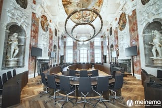 Luxe barokvergaderruimte met ronde houten vergadertafel, zwarte ergonomische bureaustoelen, witte stucplafond, rode wandschil