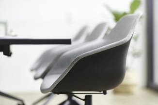 Grijze bureaustoel met ergonomische zitting en wielbasis in kantooromgeving met raam en groene plant op achtergrond.