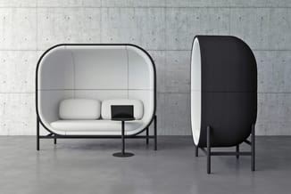Twee akoestische pods in minimalistische setting: links witte pod met zitgelegenheid en tafeltje, rechts zwarte gesloten pod.