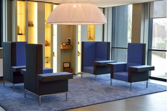 Moderne kantoorlounge met blauwe en zwarte modulaire zitmeubels, hangende roze koepellamp, gele verlichte wandkasten en grijs