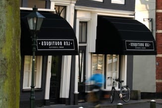 Straatgevel met twee zwarte luifels boven winkeletalages, fiets geparkeerd, bakstenen gevel met witte raamomlijsting.