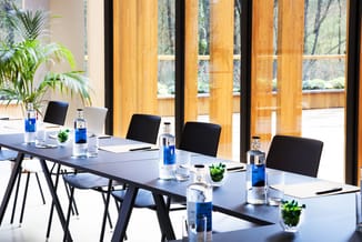 Moderne vergaderruimte met lange witte tafel, zwarte stoelen, laptops, waterflessen en groene plantjes. Grote ramen met houte