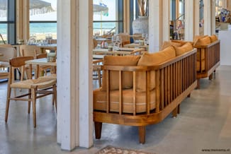 Loungeset met houten frame en cognackleurig leder in beachclub-setting met grote ramen, uitzicht op zee en naturelle houten s