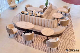 Lounge-opstelling met beige gestoffeerde bank, loungestoelen, ronde houten tafels op golvend tapijt in modern kantoor.