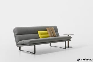 Grijze design bank met zwart metalen frame, geel en beige kussen, en glazen bijzettafel, tegen witte achtergrond.