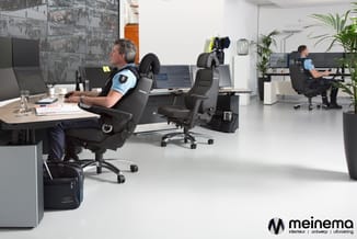 Modern kantoor met zwarte gaming-stoelen, bureaus met monitoren, planten en grote ramen. Meinema logo rechtsonder.