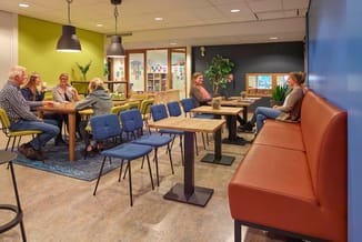 Moderne kantoorruimte met oranje bank, blauwe stoelen, houten tafels, gele en grijze wandpanelen, hanglamp en groene planten.