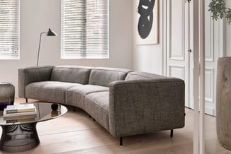 Grijze gebogen bank met textielstof en zwarte poten in moderne kantoorinterieur met ronde glazen tafel.