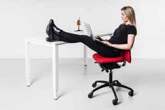 Vrouw zittend op rode bureaustoel met laptop aan wit bureau, voeten op tafelblad, kantoorscène.