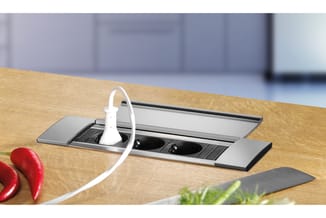 Inbouw stroomvoorziening in houten tafelblad met witte kabel en twee zwarte stopcontacten, decoratief afgewerkt.