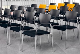 Vergaderzaal met rijen zwarte kantoorstoel met armleuningen en metalen frame. Enkele gele stoelen op achtergrond. Glazen wand