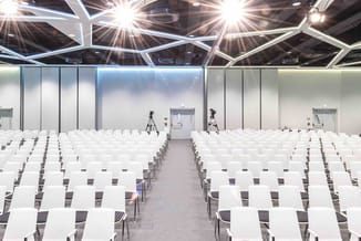 Grote vergaderzaal met witte stoelen in rijen, geometrisch plafond met LED-verlichting, grijze vloer en witte wanden.