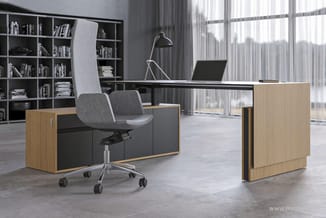 Modern kantoor met houten U-bureaus, grijze bureaustoel, zwarte lamp, en zwarte boekenkasten tegen ramen met gordijnen.