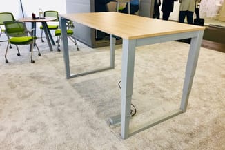 Hoge tafel met licht houten blad en grijs metalen frame, staand in kantoorruimte met grijze vloerbedekking en groene stoelen