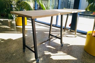 Houten tafel met zwart metalen frame en wielen in moderne kantoorruimte met groene planten en gele accent-objecten.
