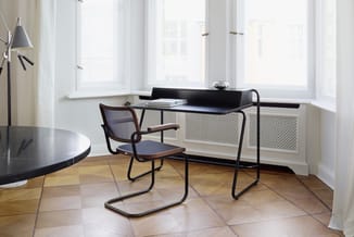 Kantoorwerkplek met zwart bureau, bruine bureaustoel en staande lamp bij ramen met radiatorombouw.