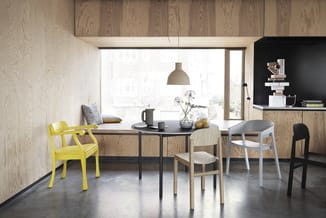 Eetkamer met zwarte tafel, gele stoel, houten stoelen, zitbank bij raam, hanglamp, houten wanden en betonvloer.