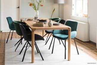 Houten eettafel met zes stoelen in groen en zwart fluweel, op beige vloerkleed in lichte kamer met bloemen en raam.