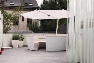 Modern terrasmeubel: witte loungeset met hoekbank, bijzettafel en parasol op houten terras naast glazen afscheiding.