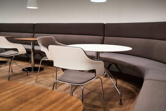 Modern kantoorlounge met grijze zitbank, witte ergonomische stoelen met grijze zitting, witte tafel en houten vloer.