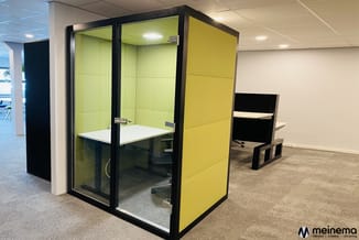 Gele belcel met zwart frame en glazen deuren in modern kantoor, voorzien van witte tafel en bureaustoel.