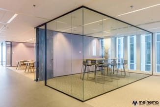 Glazen vergaderruimte met zwart frame in modern kantoor, ingericht met hoge tafel, barkrukken en gele vaas. Aangrenzende ruim