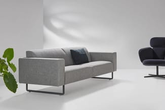 Grijze loungebank met zwart metalen onderstel en donker kussen, naast zwarte fauteuil in minimalistische kantoorruimte met pl
