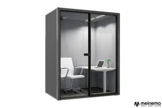 Gesloten belcel met glazen deuren en zwart frame, grijze akoestische wanden, wit bureau, bureaustoel en laptop, groene plant.