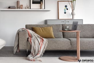Grijze stofsofa met geel kussen en geruit plaid, houten bijzettafel met laptop, wandplank met decoratie en grijs sideboard op