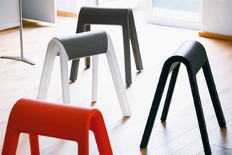 Vier stapelstoelen in verschillende kleuren (rood, wit, grijs, zwart) met gepolsterde zitting, opgesteld op houten vloer in k