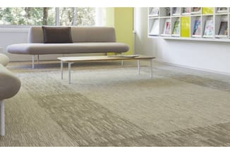 Modern kantoor met grijze ronde bank, beige tafel, grijsgroen tapijt, witte wandkasten en geel accent aan muur.