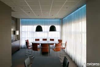 Moderne vergaderruimte met ronde witte tafel, cognackleurige stoelen, zwarte hanglampen en grote raampartijen met verticale l