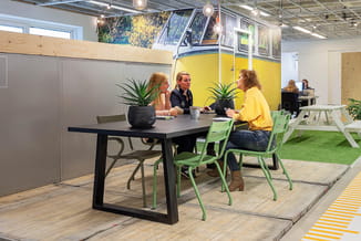 Moderne kantoorruimte met zwarte tafel, groene stoelen en gele VW-busbank tegen grijze wand. Groen kunstgras en houten vloer