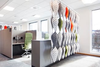 Modern kantoor met grijze akoestische scheidingswand voorzien van witte en gekleurde bladmotieven, groen gestoffeerde bureaus